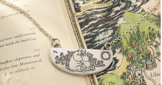 Moomin jewellery arranged in a warm summer setting with flowers, a book and friendship-inspired details