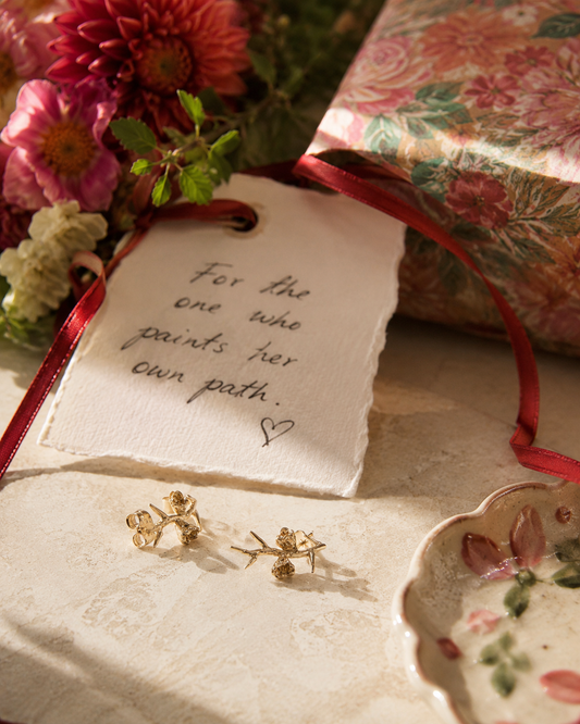 A styled flat-lay featuring a pair of gold floral stud earrings on a textured cream surface next to a handmade paper tag with a quote, a red ribbon, flowers, and a floral gift box.