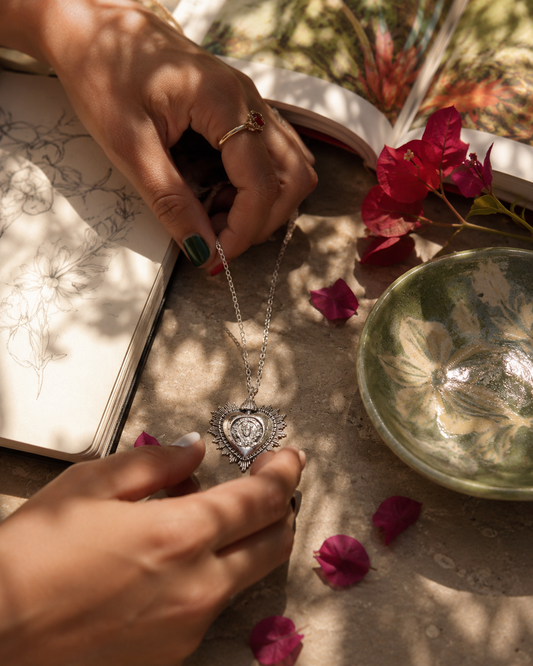 A Frida Kahlo Large Viva La Vida heart necklace in sterling silver with a central rose motif is held by hands over a styled flat-lay scene featuring an open sketchbook, red flowers, and a green glass bowl.