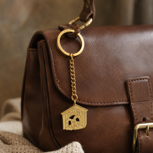A close-up of a brown leather handbag with a Wallace and gromit brass bag charm hanging shaped like a small house with a gromit dog in the centre, resting on a textured beige fabric surface.