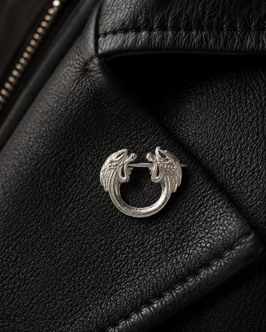Close-up of a decorative sterling silver pin brooch pinned to a black leather jacket lapel, showing engraved detail and texture.