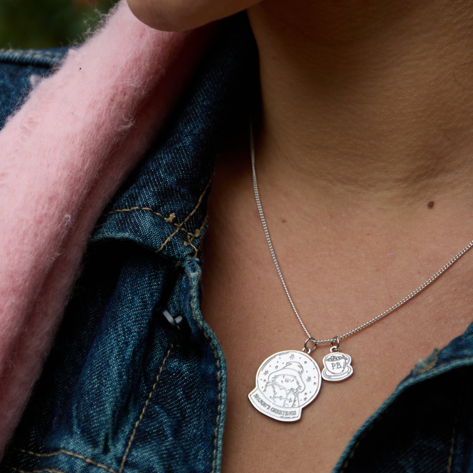 Licensed to Charm sterling silver Paddington snow globe necklace worn on a model, layered with mini PB charm, shown against a denim jacket and pink scarf. Perfect Christmas gift for her
