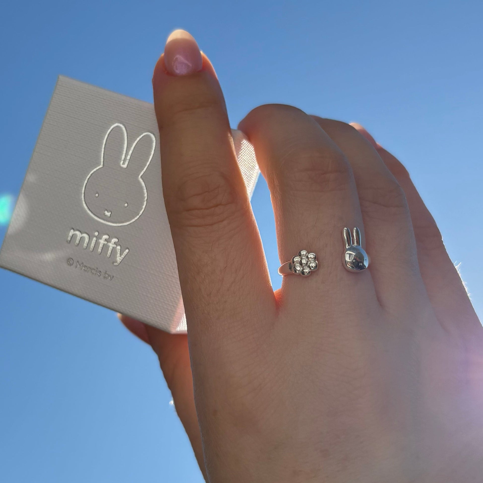 Licensed To Charm Hand wearing a sterling silver Miffy ring with a rabbit design, holding a 'miffy' branded product against a blue sky.