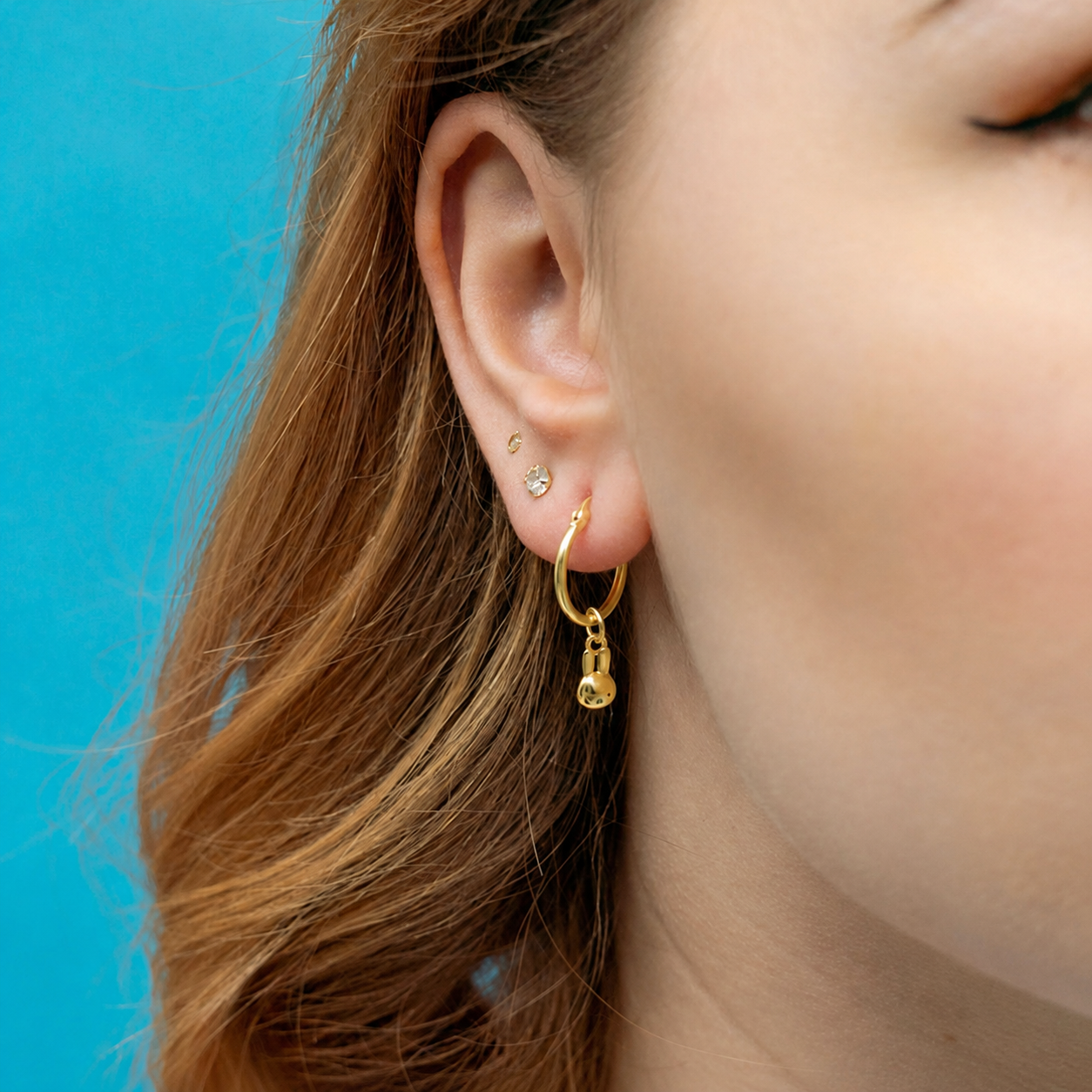 A close-up of a person's ear wearing a small Miffy  gold hoop earring with a dangling lock charm, alongside other small earrings, against a bright blue background.