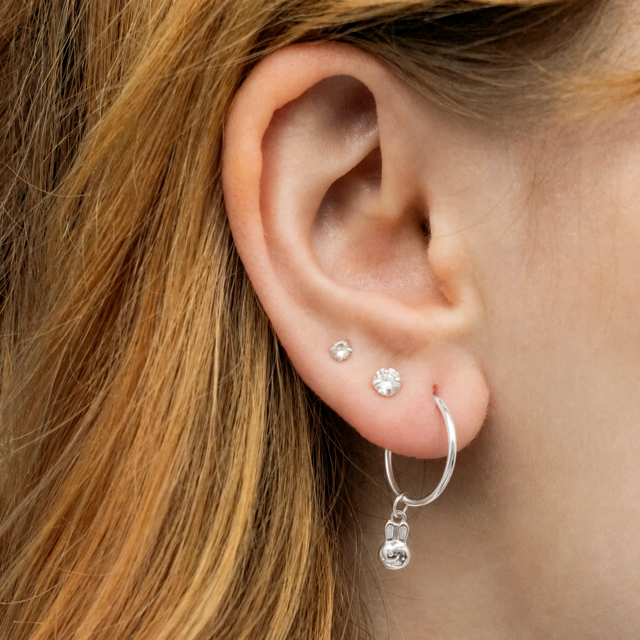 A close-up of a person's ear showcasing a silver miffy seamless hoop earring with a dangling miffy charm, along with small stud earrings.