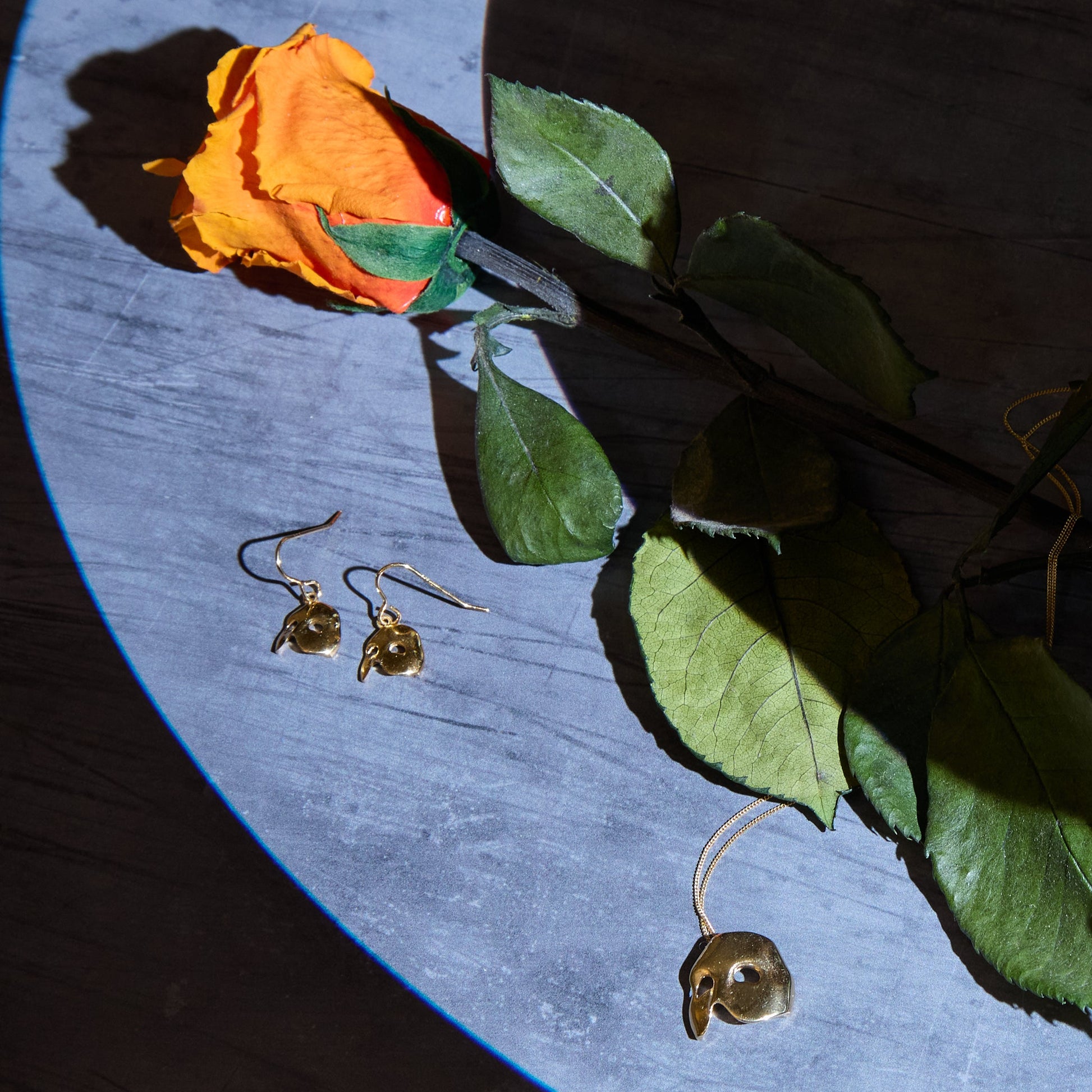 A styled flat-lay of gold phantom of the opera jewellery, including a mask necklace, a pair of dangling mask earrings, and a mask bracelet, arranged on a gray surface alongside a rose stem with green leaves and an orange rose under dramatic spotlighting.