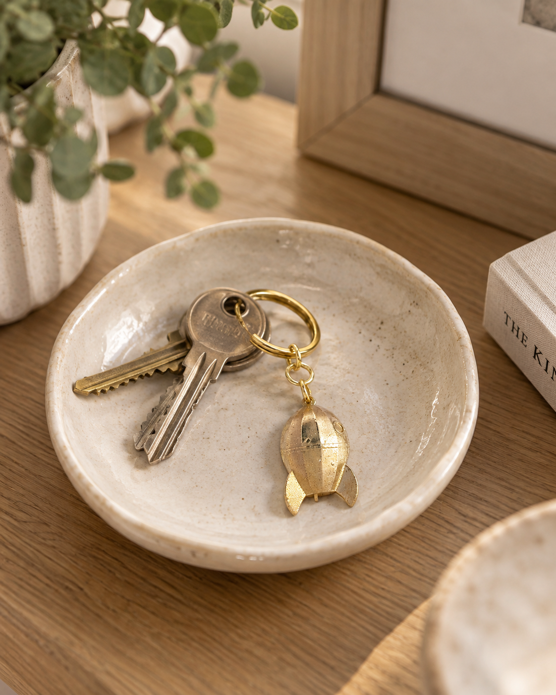 A small round ceramic key holder dish on a wooden table, holding a Wallace & Gromit Rocket brass keyring. A potted plant, framed picture, and book are softly blurred in the background.