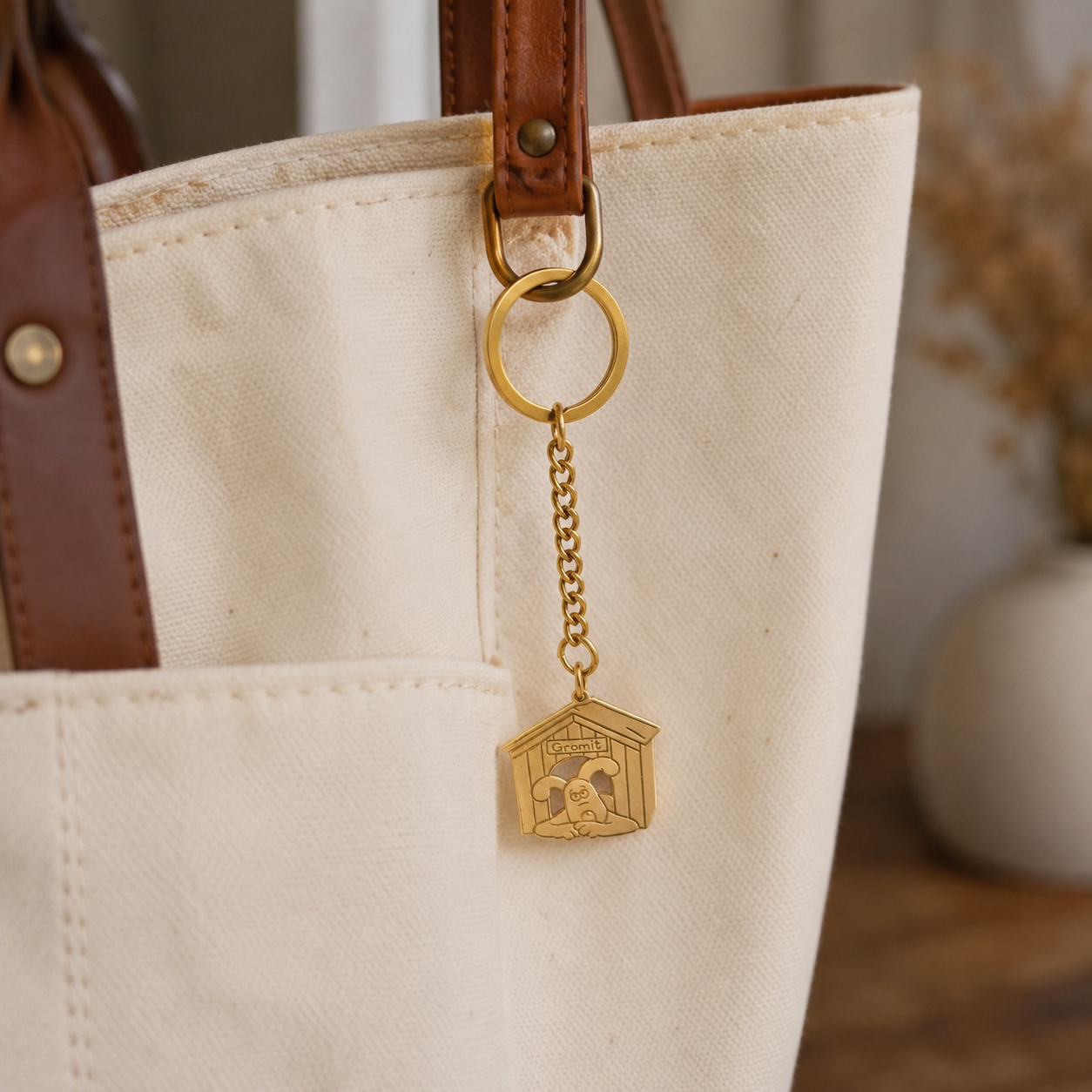 A brass metal keychain with a dog house-shaped charm with Gromit engraved, hanging from a brown leather strap of a canvas tote bag.