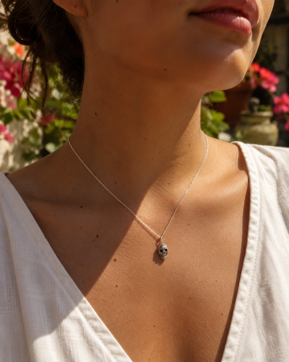 Frida Kahlo Aztec Skull Necklace in sterling silver worn with a cream blouse in warm sunlight, styled against a blurred garden background with pink flowers.