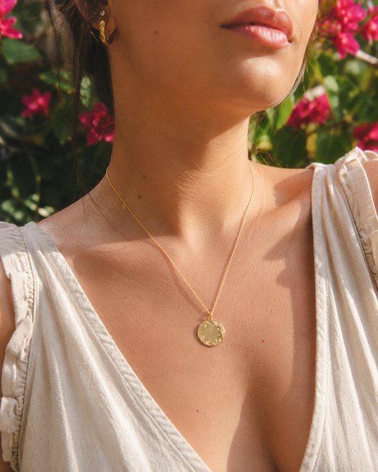 Frida Kahlo Folk Disc Necklace in 18ct gold vermeil worn with a cream top in warm sunlight, styled against a blurred garden background with pink flowers.