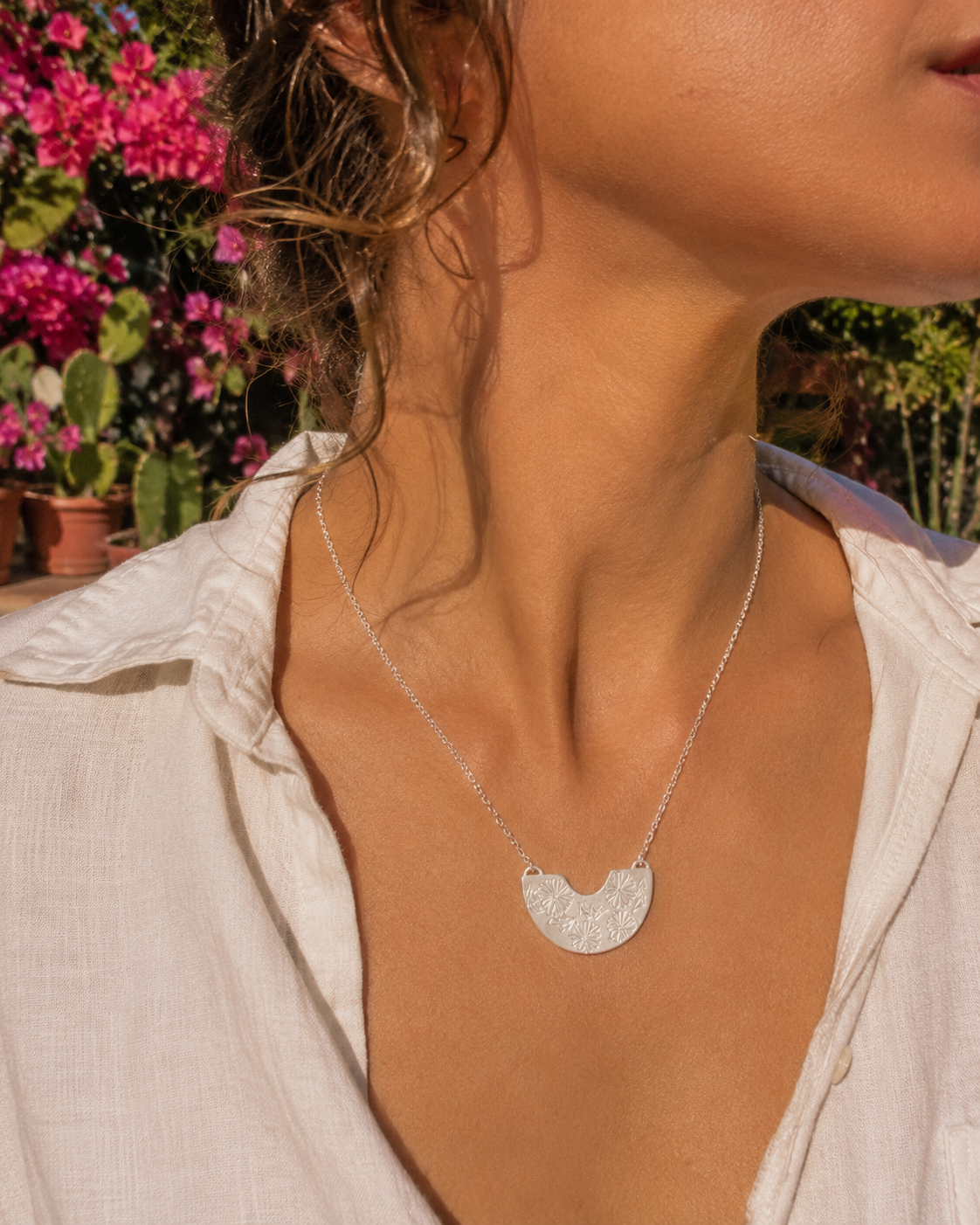 A close-up lifestyle shot of a woman wearing a Frida Kahlo silver crescent moon pendant necklace with a textured, hammered finish. She is wearing a white linen shirt, with a sunny outdoor garden background featuring pink flowers.