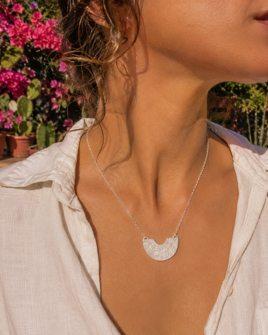A close-up lifestyle shot of a woman wearing a Frida Kahlo silver crescent moon pendant necklace with a textured, hammered finish. She is wearing a white linen shirt, with a sunny outdoor garden background featuring pink flowers.