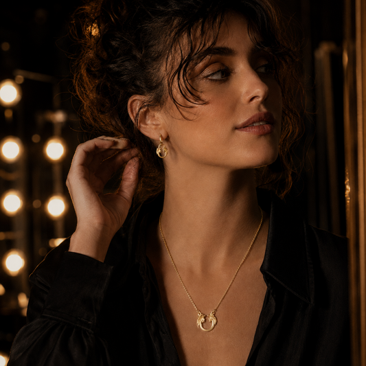 Model wearing the Jesus Christ Superstar gold logo necklace and matching earrings in a warm theatre-inspired lifestyle image.