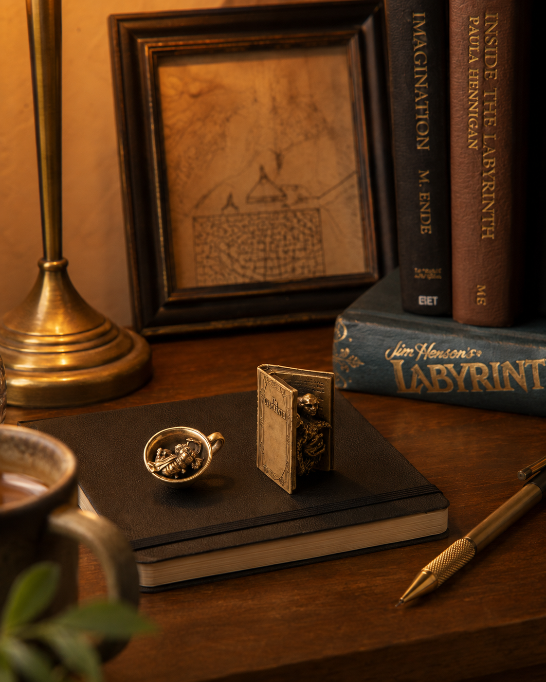 Labyrinth Character Book and Worm in a Cup miniature bronzes styled on a warm collector’s desk with books and a sketchbook.