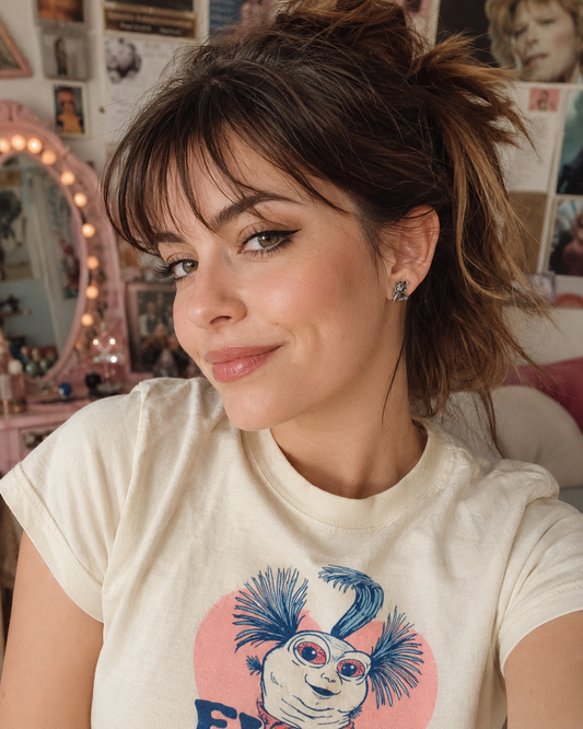 Woman wearing small Labyrinth Worm stud earrings and a cream Worm T-shirt in a nostalgic 80s-inspired bedroom.
