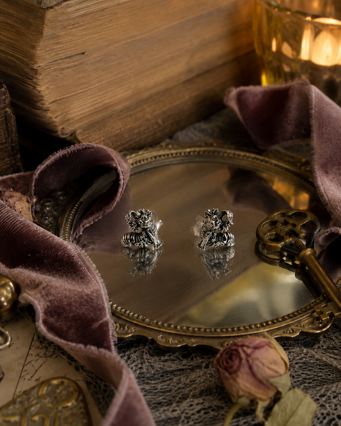 A pair of silver labyrinth worm stud earrings displayed on a decorative oval mirror, surrounded by vintage props including an antique key, a dried rose, purple velvet fabric, and old books.