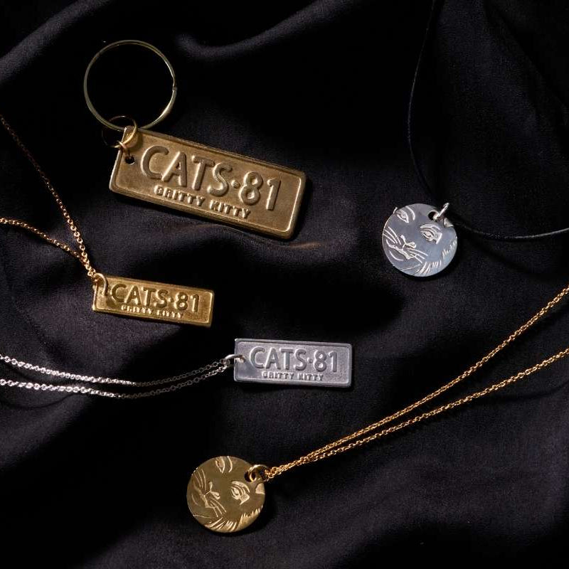 Licensed To Charm CATS jewellery collection featuring gold and silver necklaces, a CATS 81 keyring and character pendant necklaces on black fabric