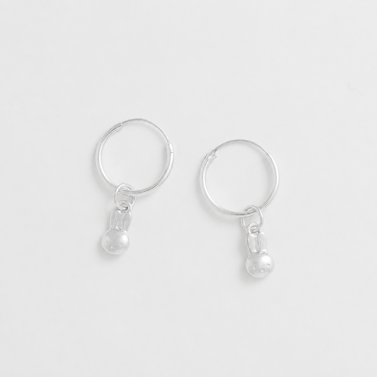 A pair of silver seamless hoop earrings with small dangling miffy charm, shown on a clean light gray background.