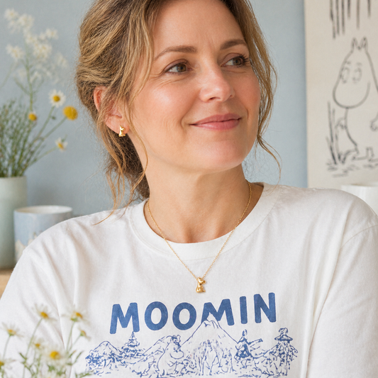 Woman wearing Licensed To Charm Moomin gold necklace and matching stud earrings, styled with a white Moomin t-shirt in a soft Nordic-inspired lifestyle setting