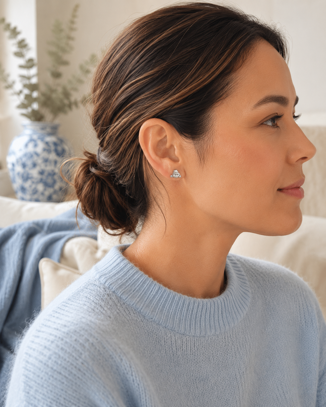 A woman in a light blue knit sweater shown in profile, wearing a small Silver Paddington stud earrings. The background features a cozy indoor setting with a blue and white patterned ceramic vase and a soft beige sofa.
