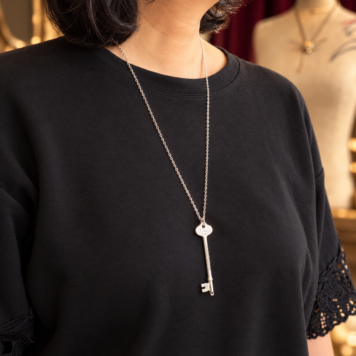 A silver phantom of the opera key pendant necklace worn by a model in a black sweatshirt, with a softly blurred boutique background.