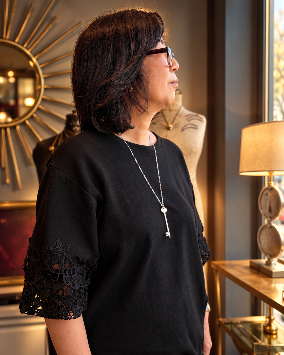 A woman with short dark hair and glasses, wearing a black short-sleeve top with lace trim on the sleeves and a silver key pendant necklace, standing in a warmly lit interior room.