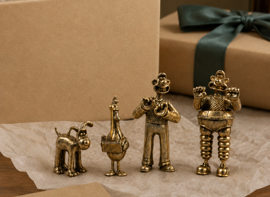 Four small bronze figurines in a gold finish, arranged from smallest to largest on brown tissue paper. The set includes a Gromit figure, Feather's McGraw figure,  Wallace figure, and wallace wrong trousers figure.