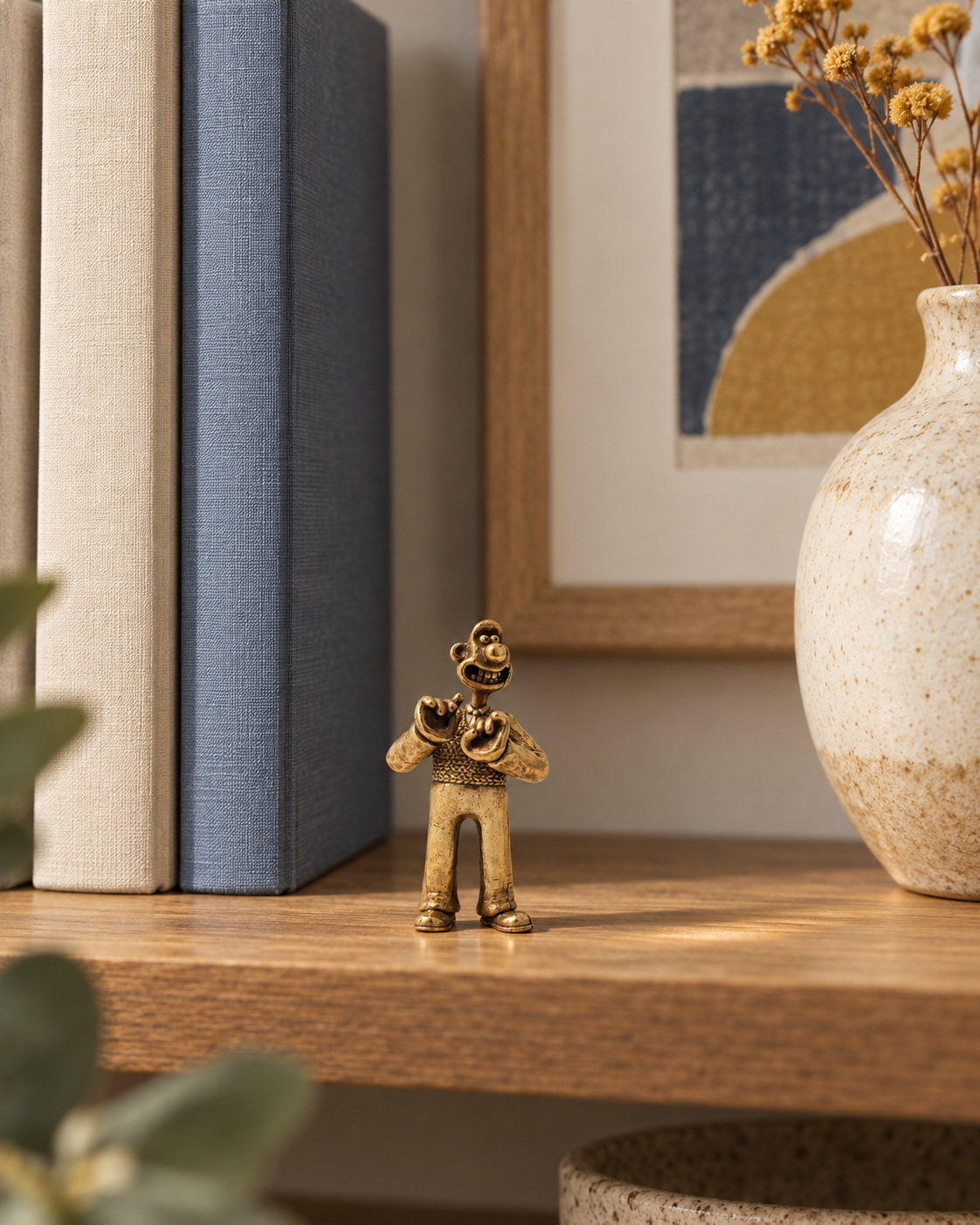Miniature Wallace bronze figurine displayed on a wooden shelf beside neutral books, a ceramic vase and a framed abstract print.
